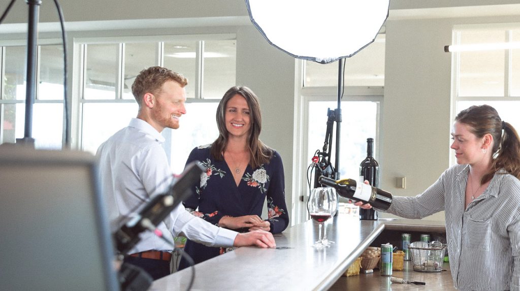 A person is pouring red wine into a glass for another person at a bar counter in a production set.
