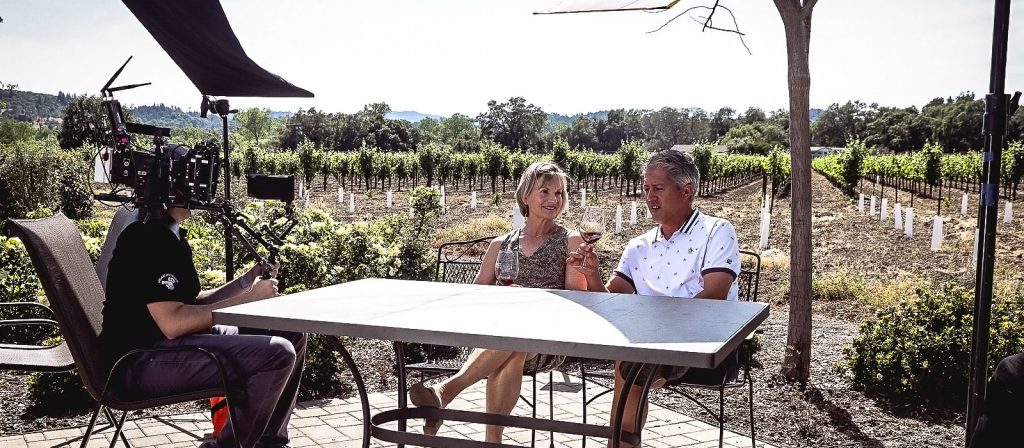 A camera crew is filming two people sitting at an outdoor table in a vineyard.
