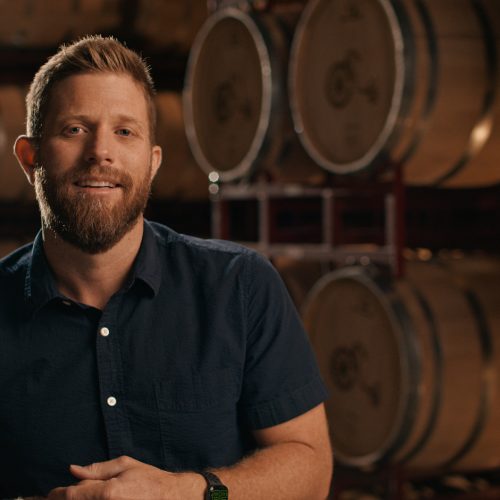 A man standing in a warehouse with rows of large wooden barrels.