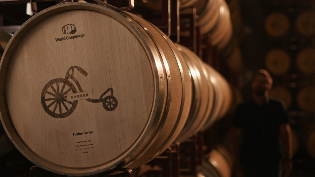 Close-up of a wooden barrel with a logo of a penny-farthing bicycle etched onto its surface, labeled HARKEN.