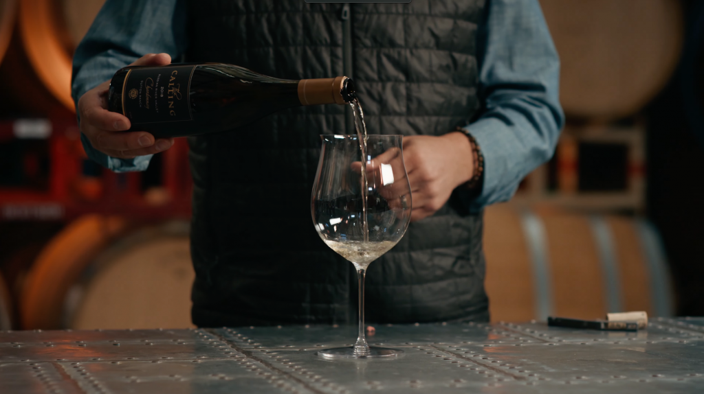 The Calling Wine Still 6 A person is pouring white wine from a bottle labeled 'The Calling' into a wine glass on a metal table.