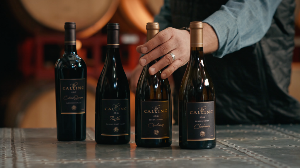 The Calling Wine Still 7 Four bottles of wine from 'The Calling' brand. A hand is holding the third bottle from the left.
