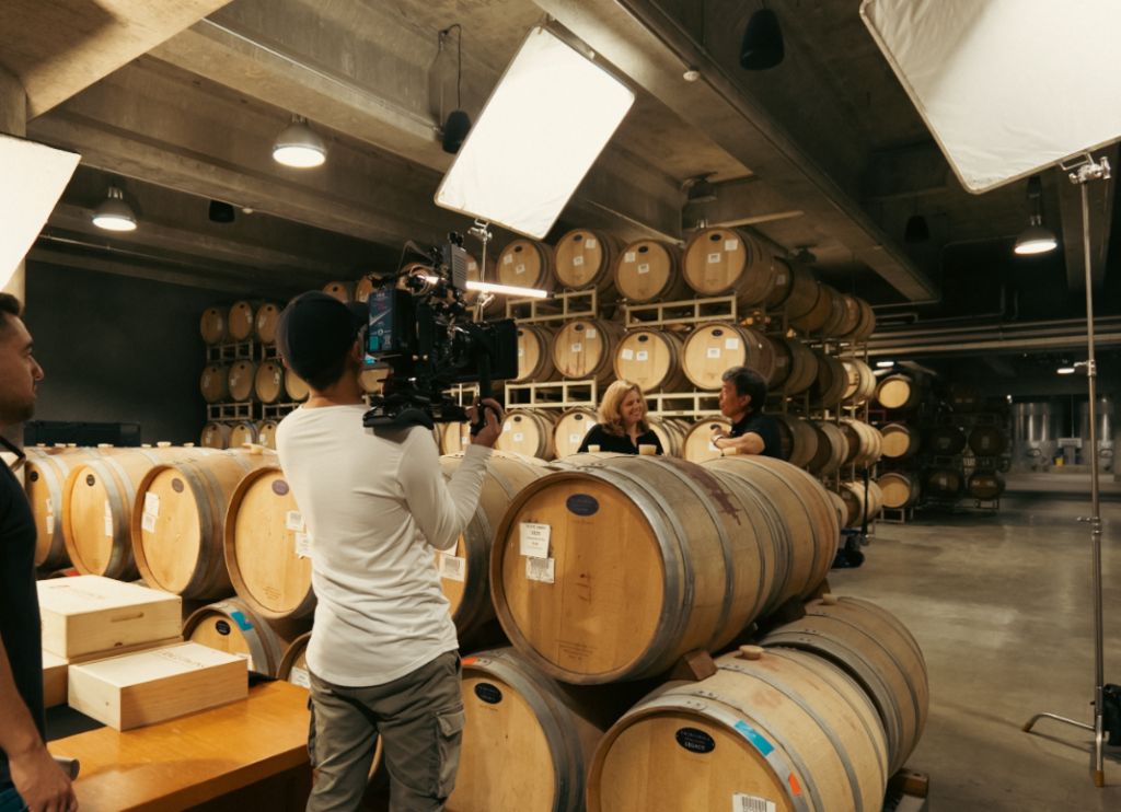 LangeTwins Winery Case Study Photo 1 A wine cellar with wooden barrels in the background. A man films two people sitting and talking near the barrels.