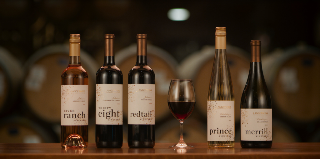 LangeTwins Winery Case Study Photo 13 Five bottles of wine labeled 'River Ranch,' 'Thirty Eight,' 'Redtail,' 'Prince,' and 'Merrill,' with a glass of red wine between the third and fourth bottles, set on a wooden surface.