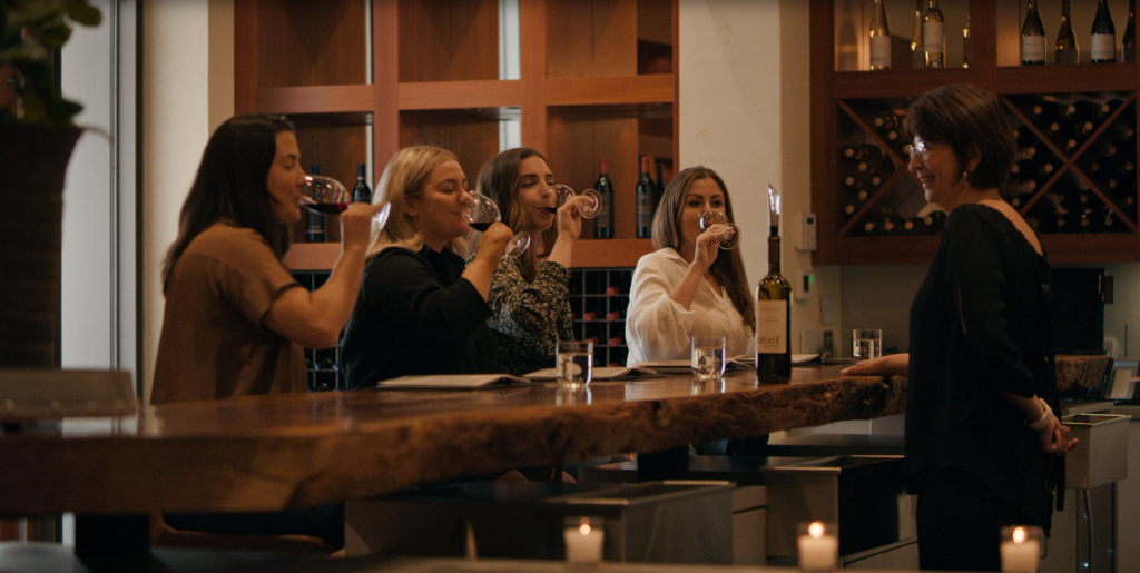 LangeTwins Winery Case Study Photo 19 Group of four women seated at a bar counter, each holding and tasting wine glasses, with a fifth woman guiding the session.