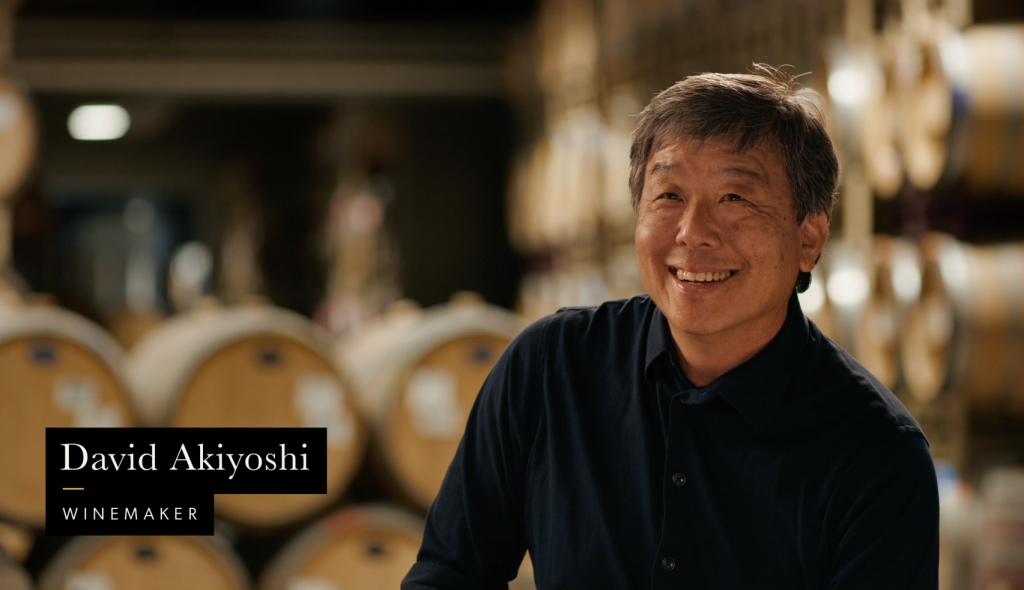 LangeTwins Winery Case Study Photo 7 A man standing in a wine cellar with wine barrels in the background and a label that reads 'David Akiyoshi, Winemaker.'